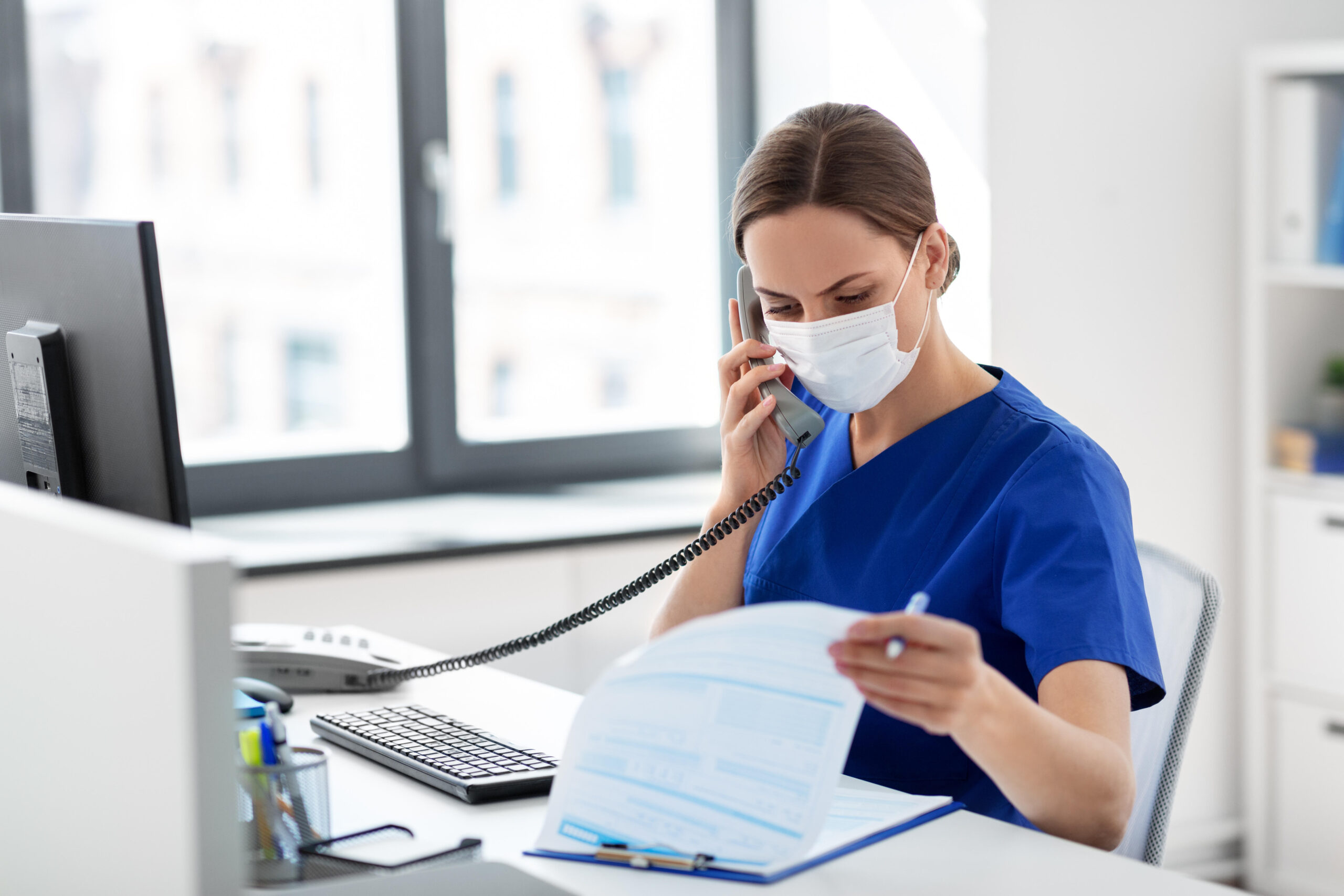 medicine, people and healthcare concept - female doctor or nurse wearing face protective medical mask for protection from virus disease with computer and clipboard calling on phone at hospital